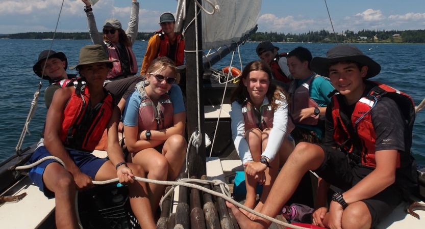 A group of students wearing life jackets sit in a sailboat and smile for the photo. 